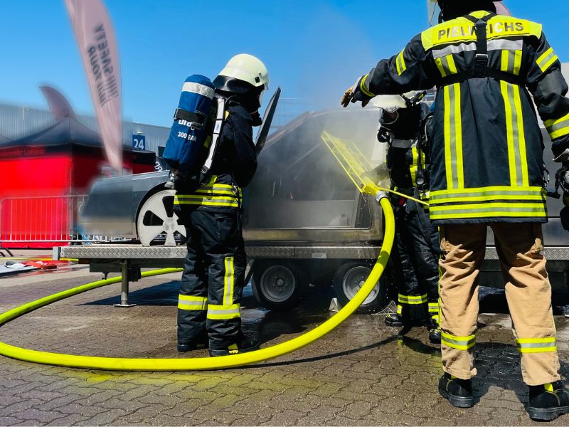 Messegelände - Auto-Attrappe aus Edelstahl - Feuerwehrleute verwenden den FIREdown SprayJet und erzeugen Sprühnebel